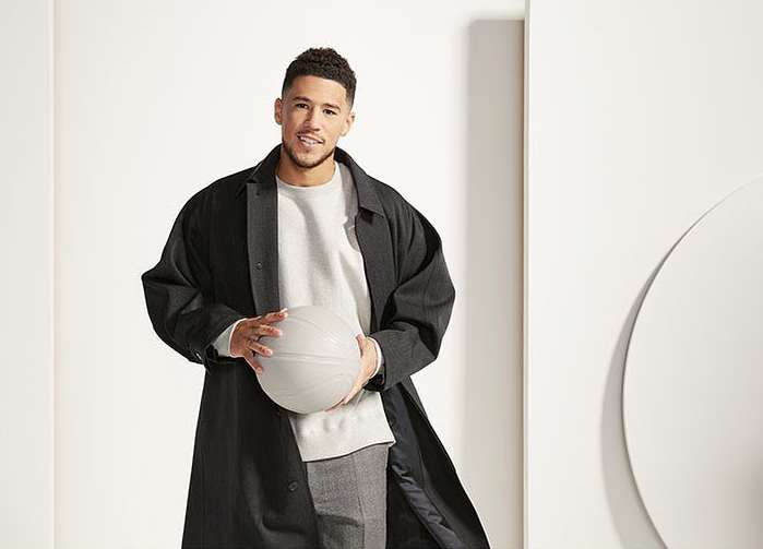 An adult male in a black coat, gray pants, and light sweater, holding a gray basketball against a white wall and background.
