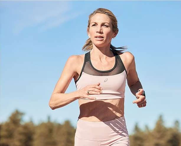 Adult woman running outdoors on a sunny day wearing a sports bra and leggings, with a clear blue sky and trees in the background.