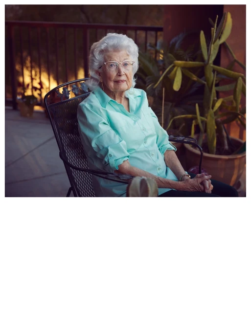 Elderly woman relaxing in chair outdoors on porch. She is wearing glasses and a light green shirt in a calm, outdoor setting.