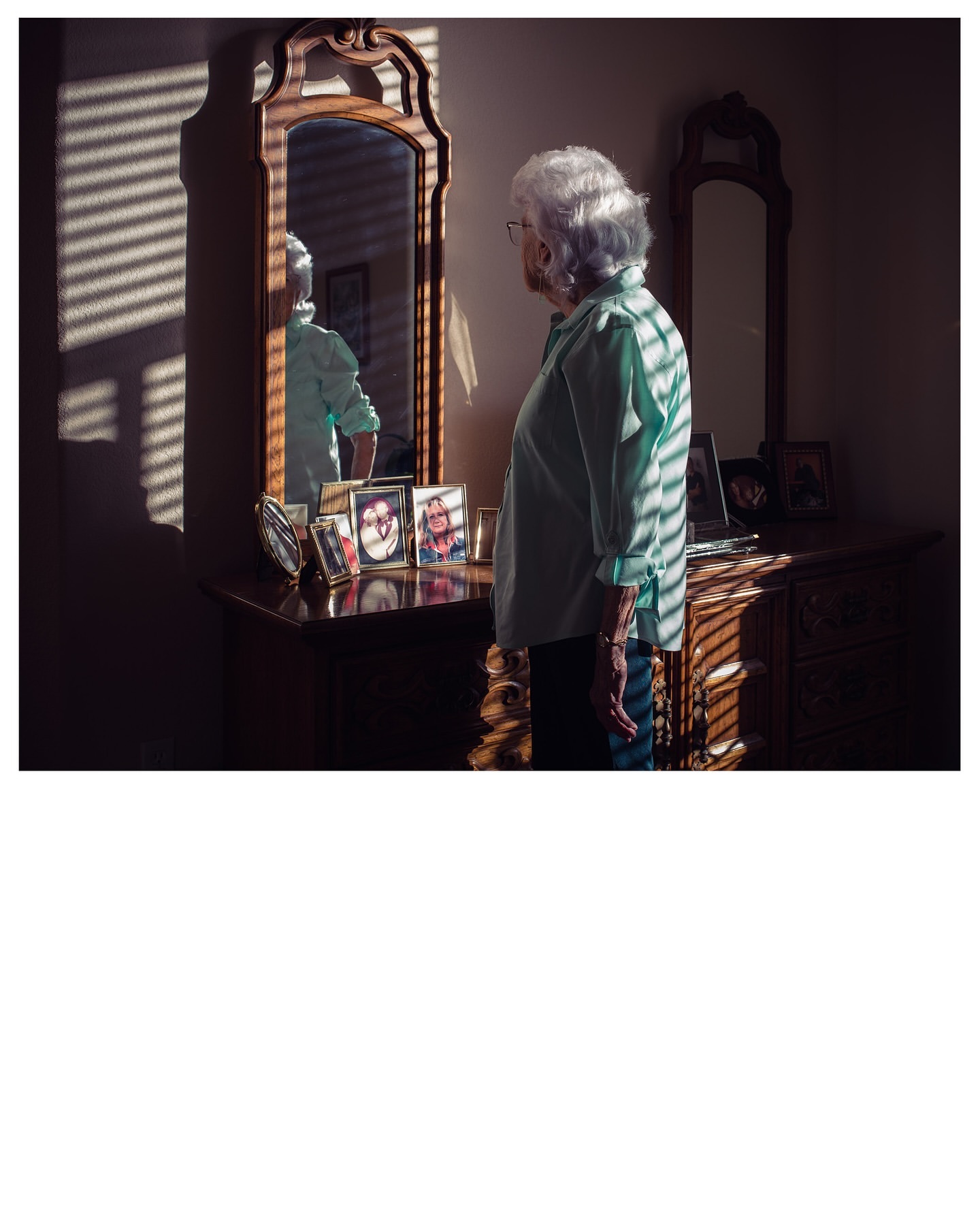 Elderly woman in a light blue shirt standing in front of a dresser with framed photos, looking at her reflection in a mirror.