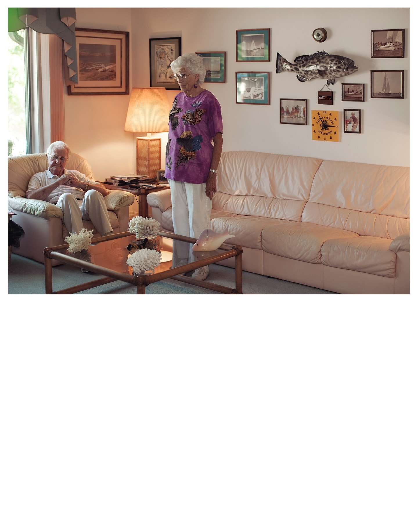 Indoor shot of an elderly couple in a living room with white walls, a couch, and framed artwork of seascapes and a mounted fish.