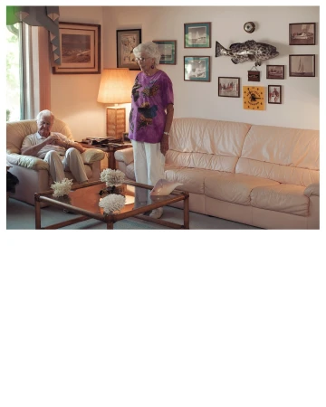 Indoor shot of an elderly couple in a living room with white walls, a couch, and framed artwork of seascapes and a mounted fish.