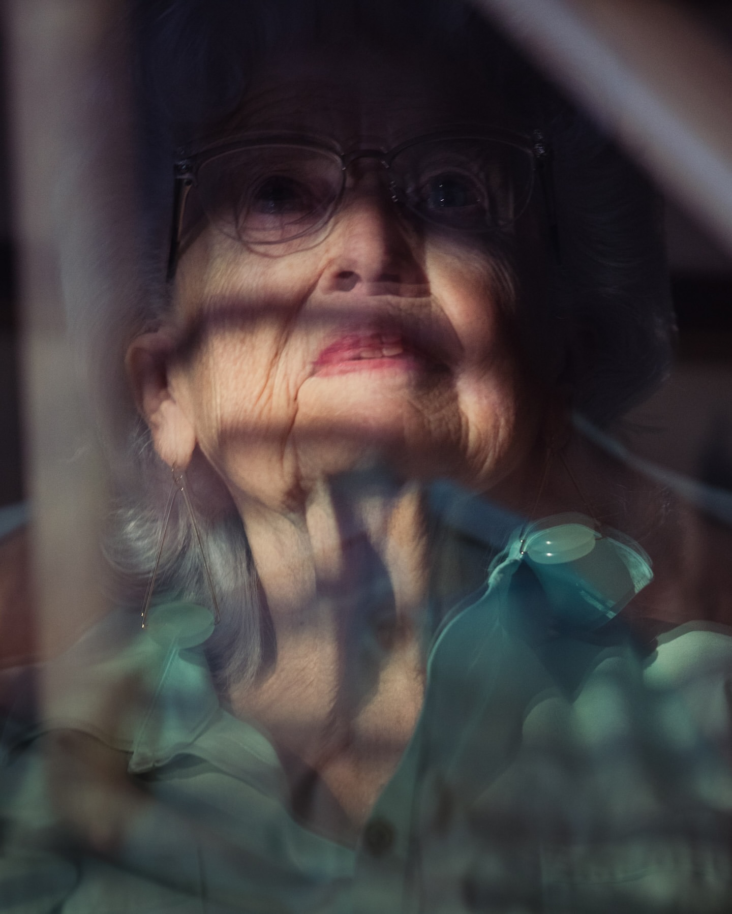 Close-up of an elderly woman wearing glasses looking through a window with sunlight creating shadows on her face and teal top.