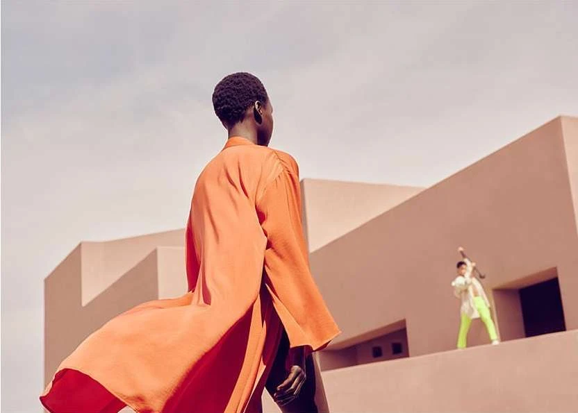 Two fashion models pose outdoors against a peach-colored architectural background, one in orange, the other in bright green.