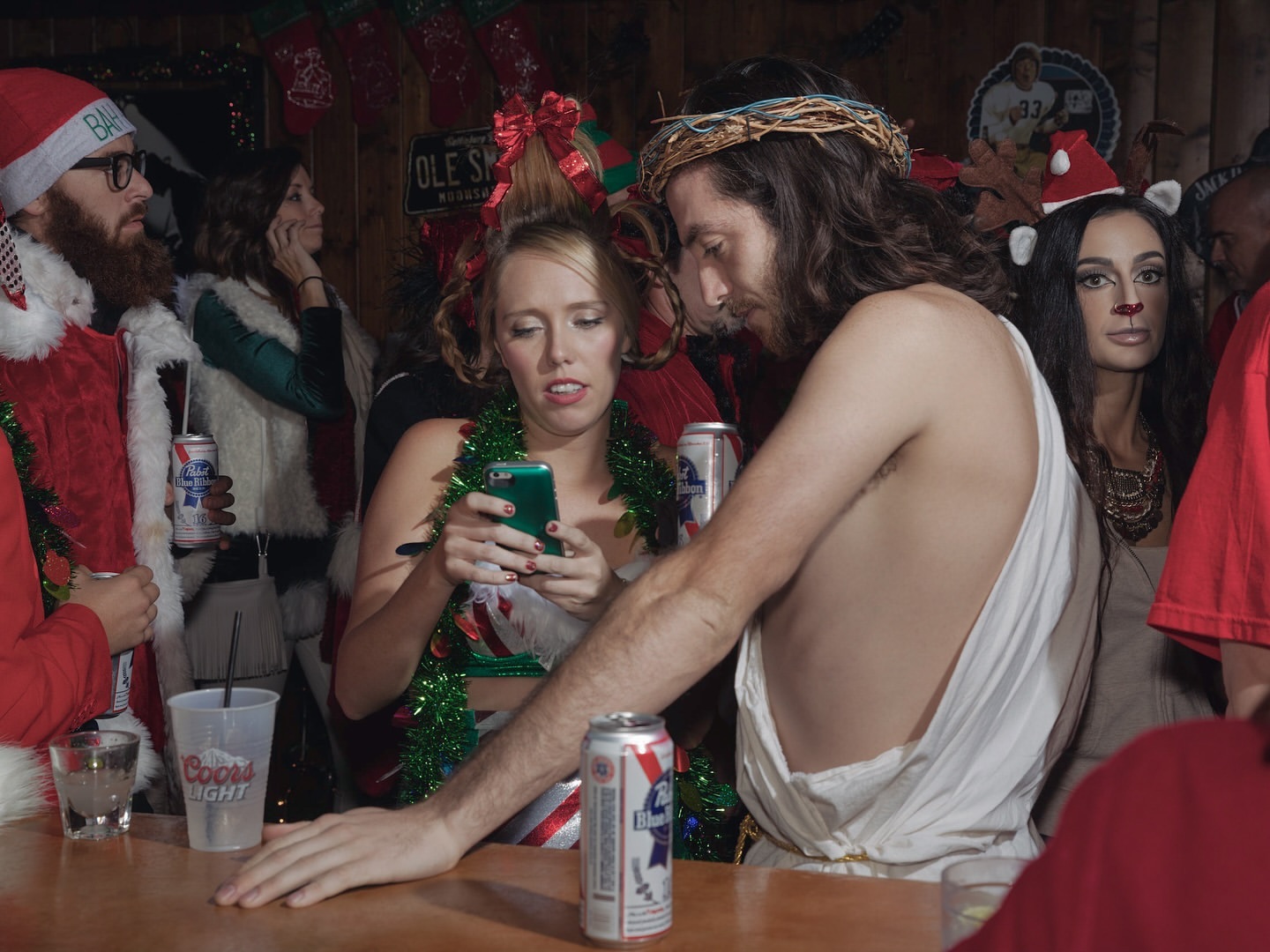 Holiday party scene with people in costumes, including Santa and Jesus, gathered at a bar with drinks and festive decorations. 