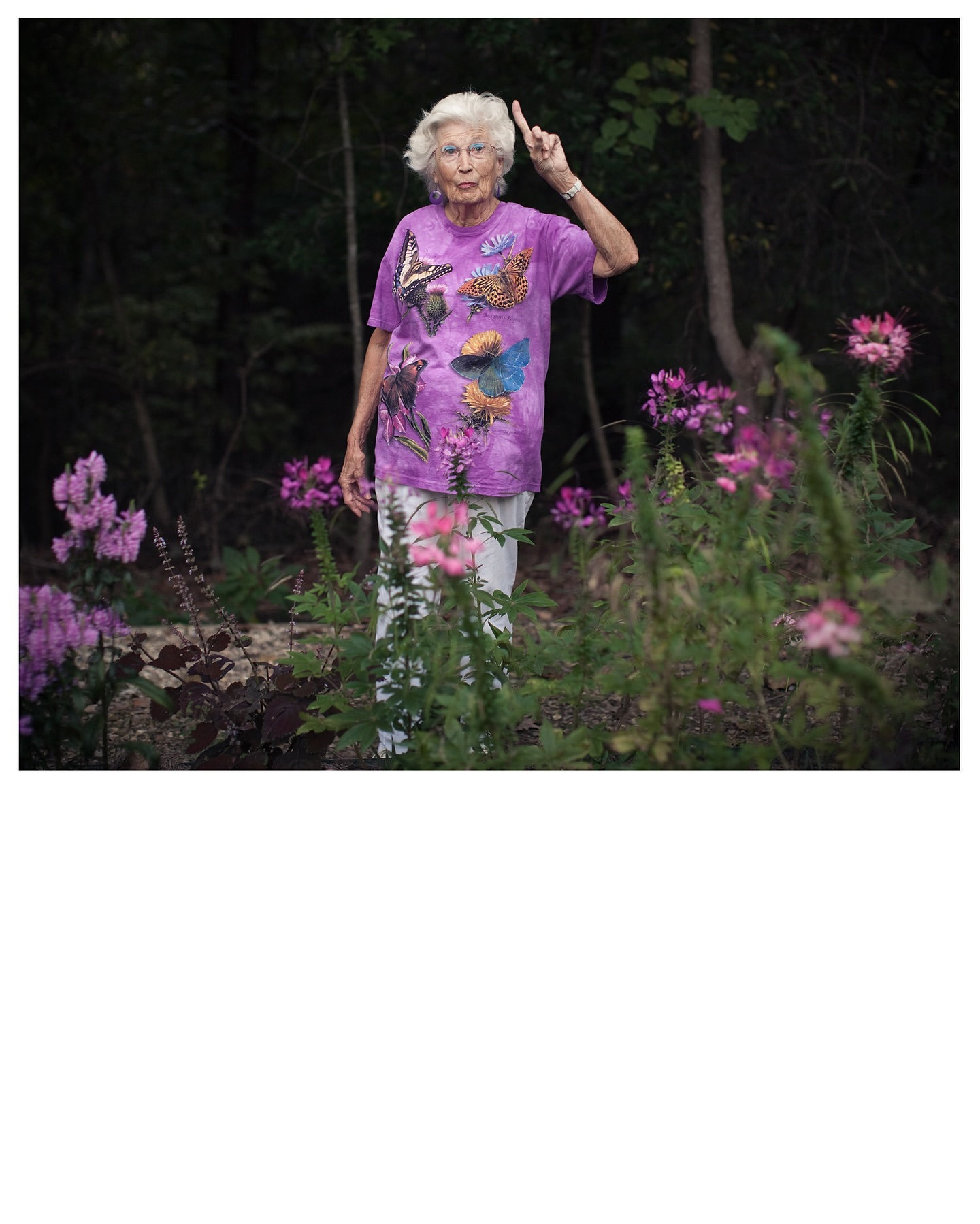 Elderly woman in a purple butterfly t-shirt stands in a garden, pointing upward with one finger raised in a lush outdoor setting.