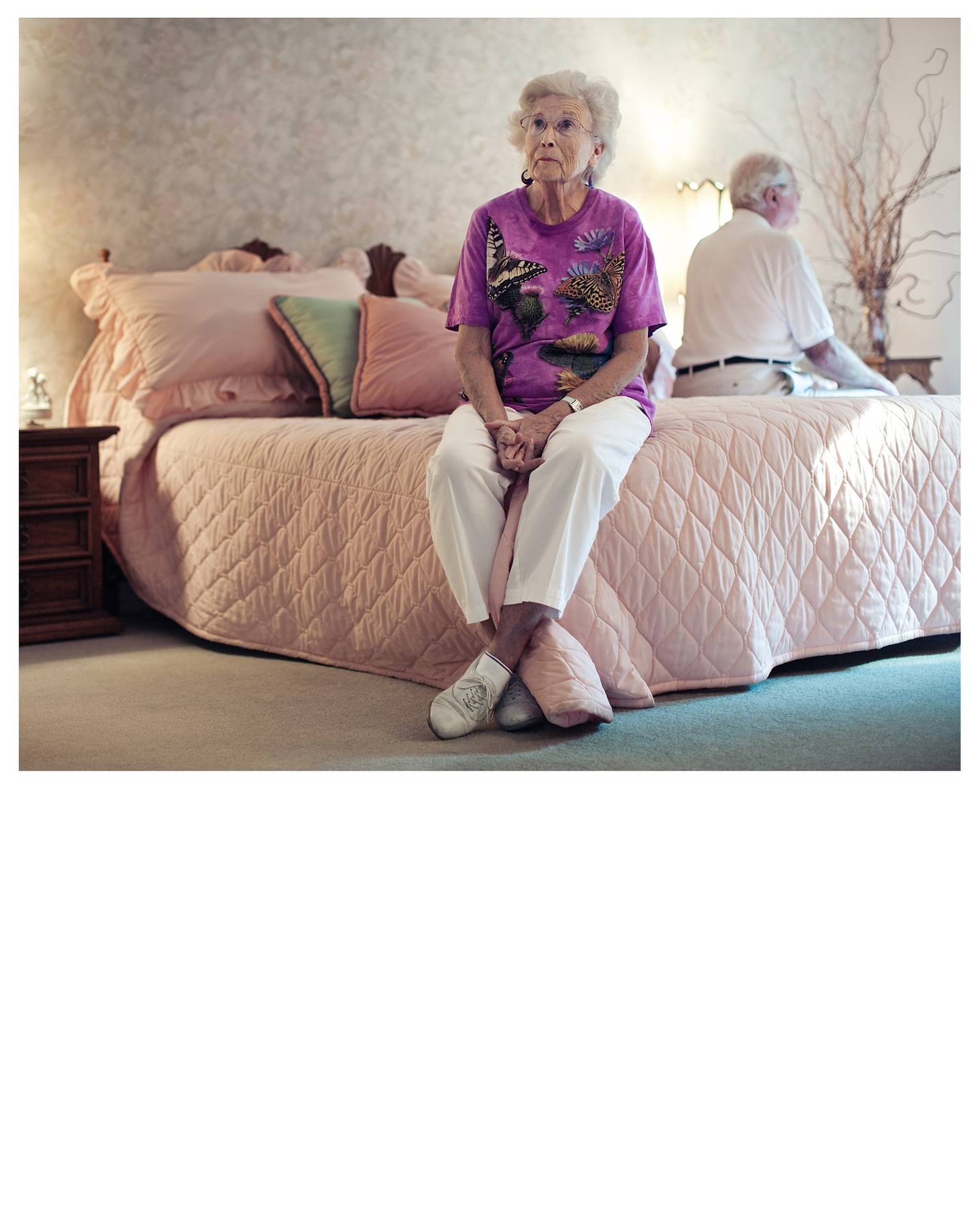 Elderly woman with short white hair sits on a pink quilted bed in a bedroom, with an elderly man in the background.