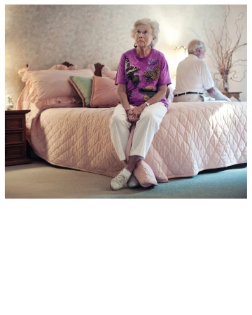 Elderly woman with short white hair sits on a pink quilted bed in a bedroom, with an elderly man in the background.