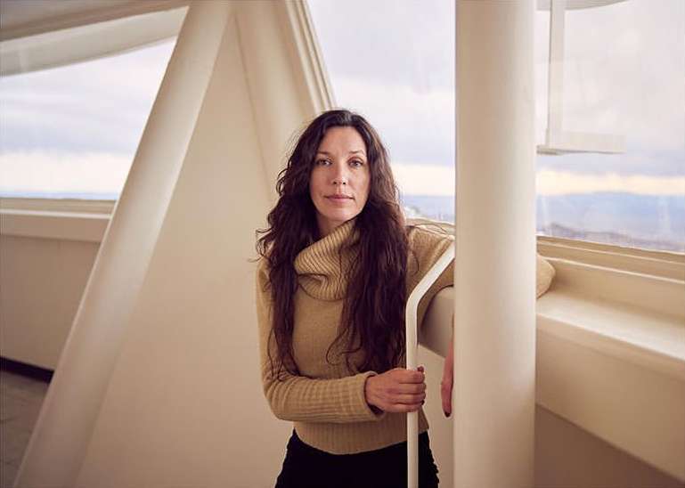 Indoor portrait of a woman with long wavy hair wearing a tan turtleneck sweater and black pants, posing in front of a window.
