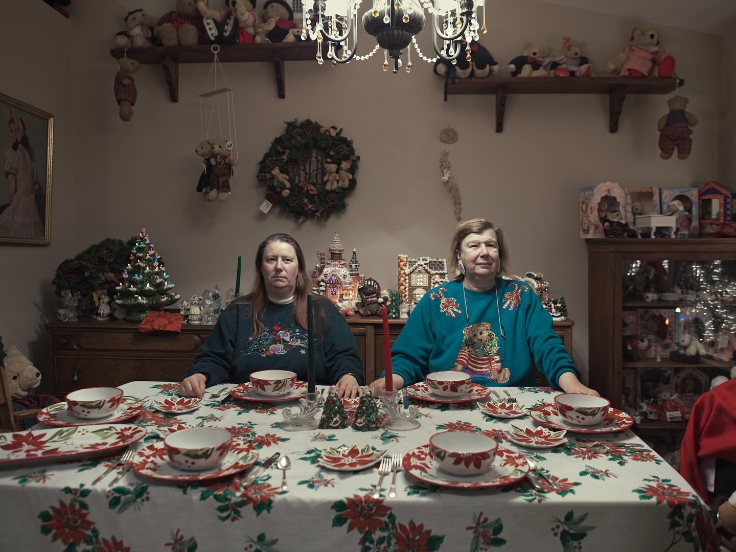 Two adult women sit at a formally set table with Christmas decor. Shelves of dolls line the background in a holiday-themed interior space.