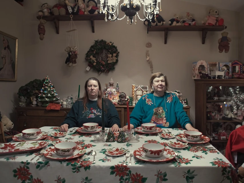Two adult women sit at a formally set table with Christmas decor. Shelves of dolls line the background in a holiday-themed interior space.
