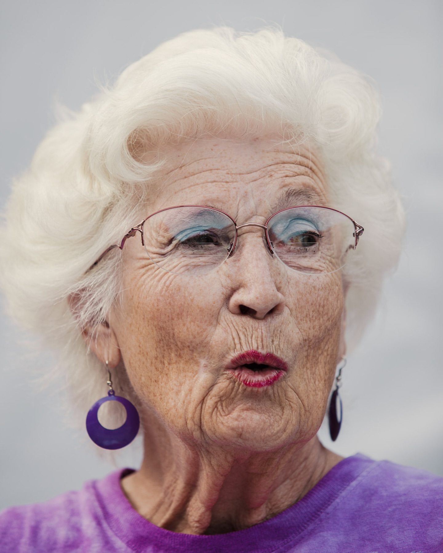 Close-up portrait of an elderly woman with white hair wearing glasses, a purple shirt and hoop earrings, with a soft neutral background.