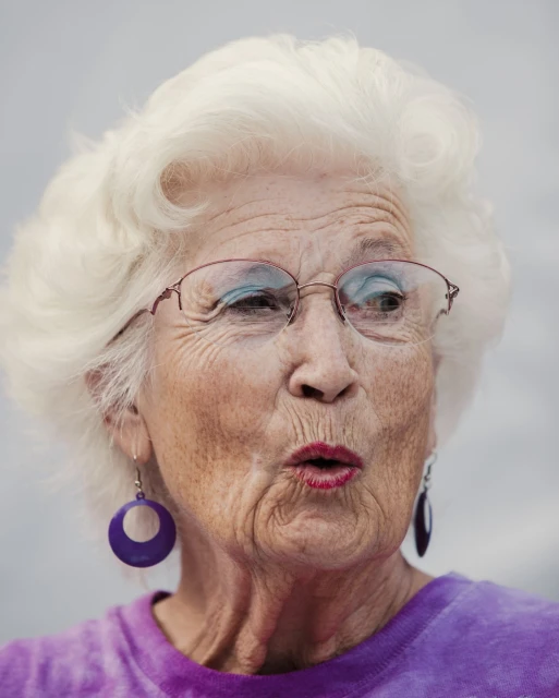 Close-up portrait of an elderly woman with white hair wearing glasses, a purple shirt and hoop earrings, with a soft neutral background.