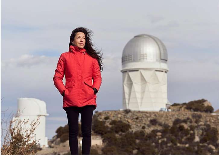 Woman in a red jacket stands near an astronomical observatory under a cloudy sky, gazing towards the horizon with wind-blown hair.