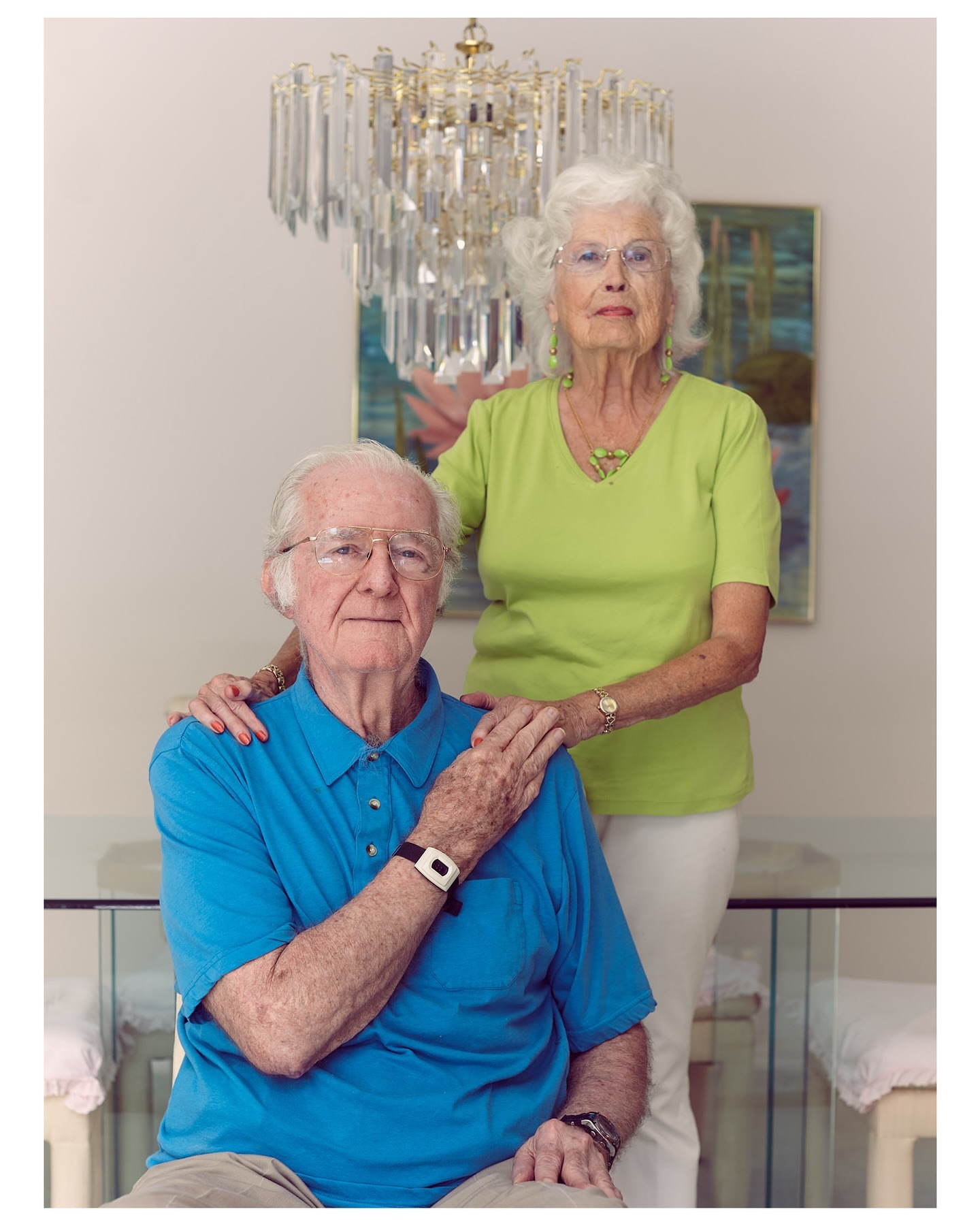 Portrait of a smiling senior couple indoors with a chandelier, painting, and dining table in the background, wearing light-colored clothing.