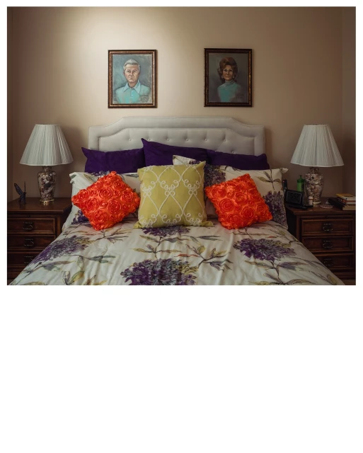 Bedroom interior featuring a bed with floral patterned bedding and pillows, two lamps on bedside tables, and portraits on the wall.