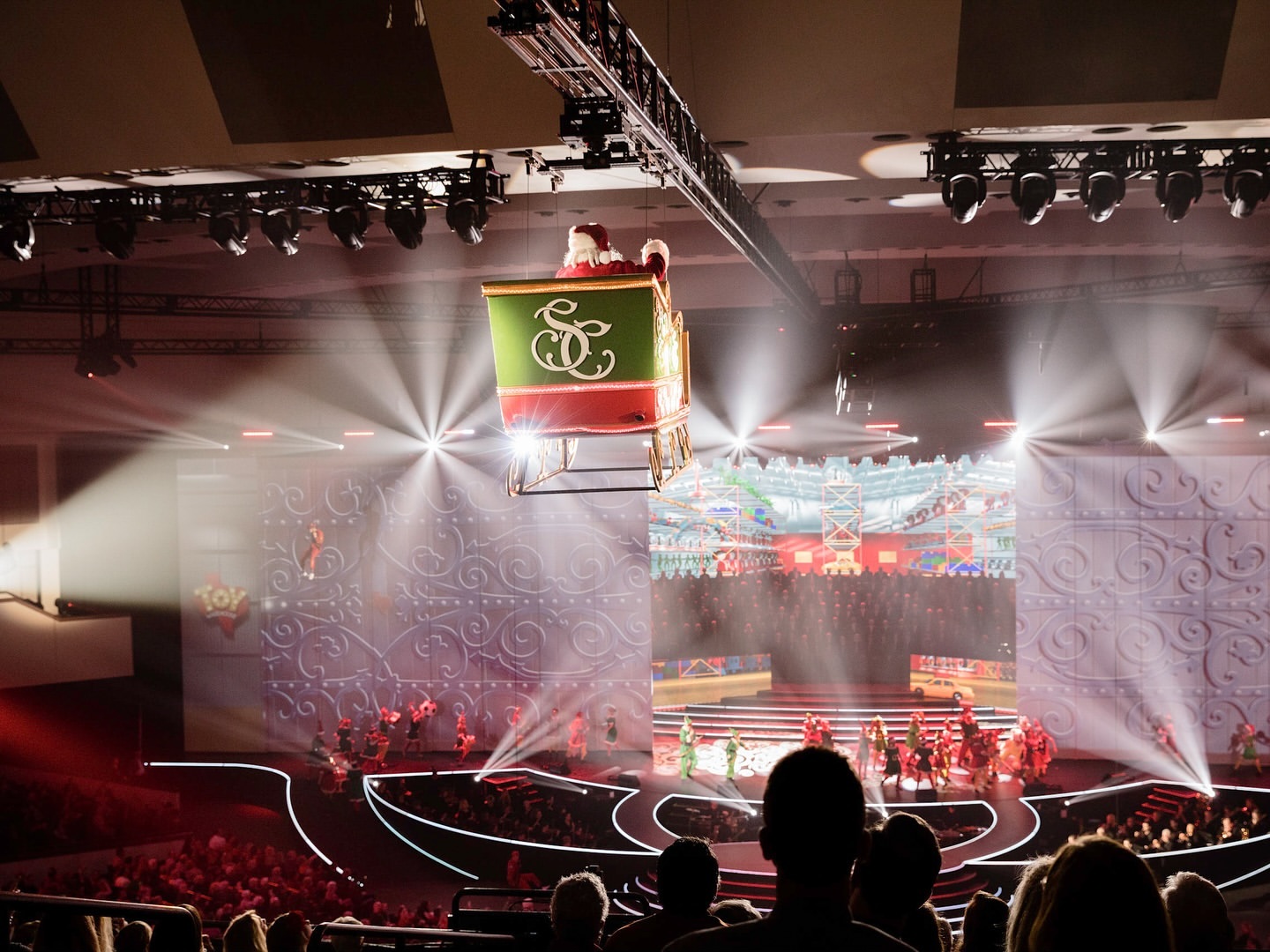 Santa in a green and red sleigh suspended above a stage with performers and a seated audience in a large auditorium space.