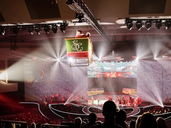 Santa in a green and red sleigh suspended above a stage with performers and a seated audience in a large auditorium space.