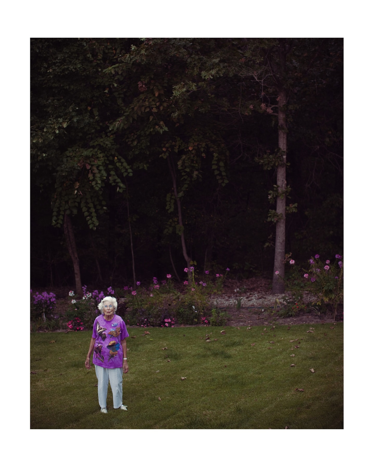 Elderly woman with white hair and purple shirt standing in a grassy backyard with trees and flowers in the background.