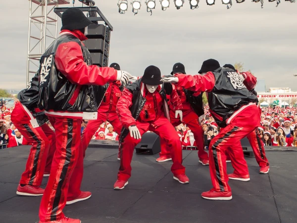 Dance troupe in red performs on outdoor stage, crowd with Santa hats visible in background; daytime event with stage lighting.