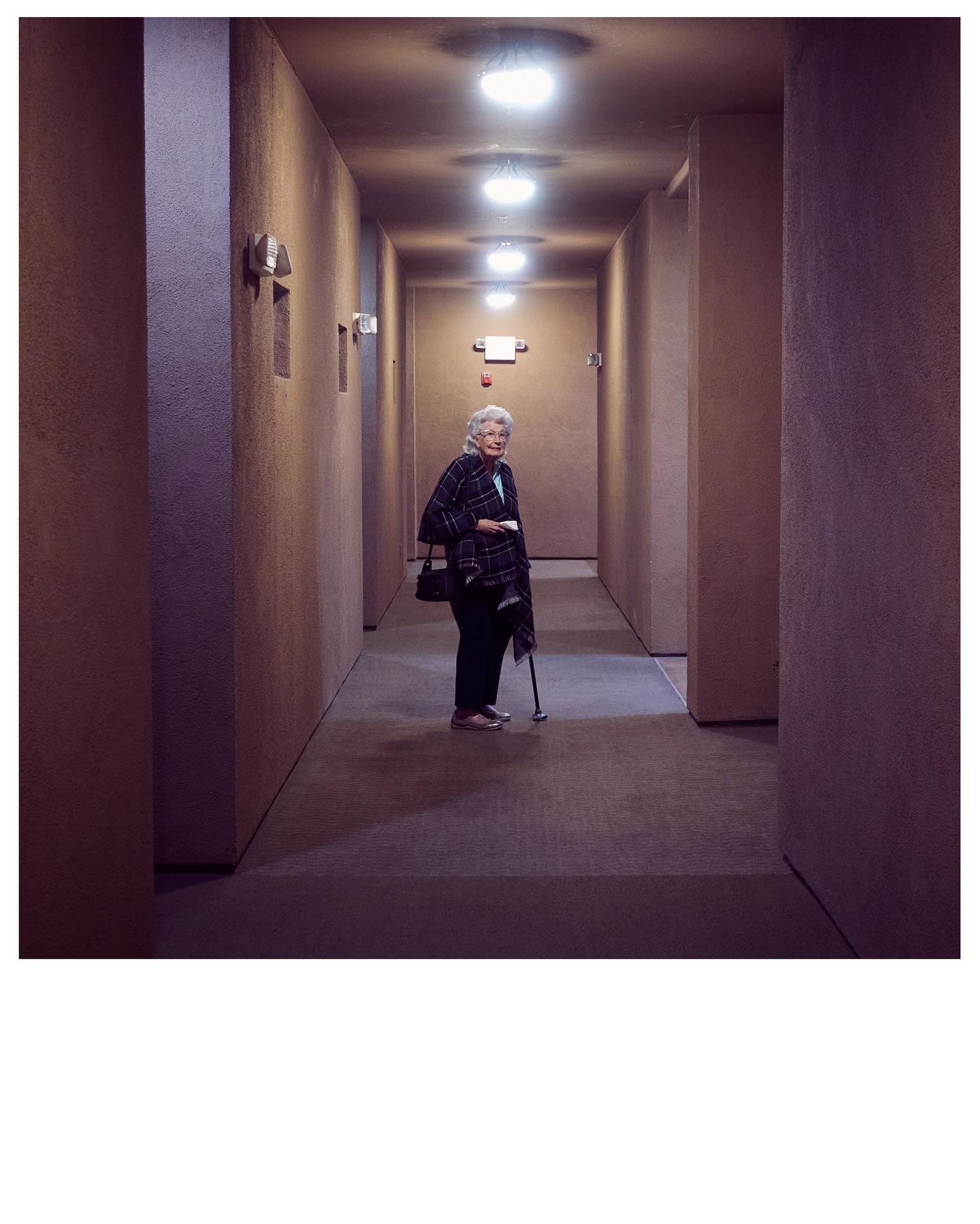 Elderly woman stands in a long hallway with lights, wearing a patterned jacket and carrying a bag and cane.