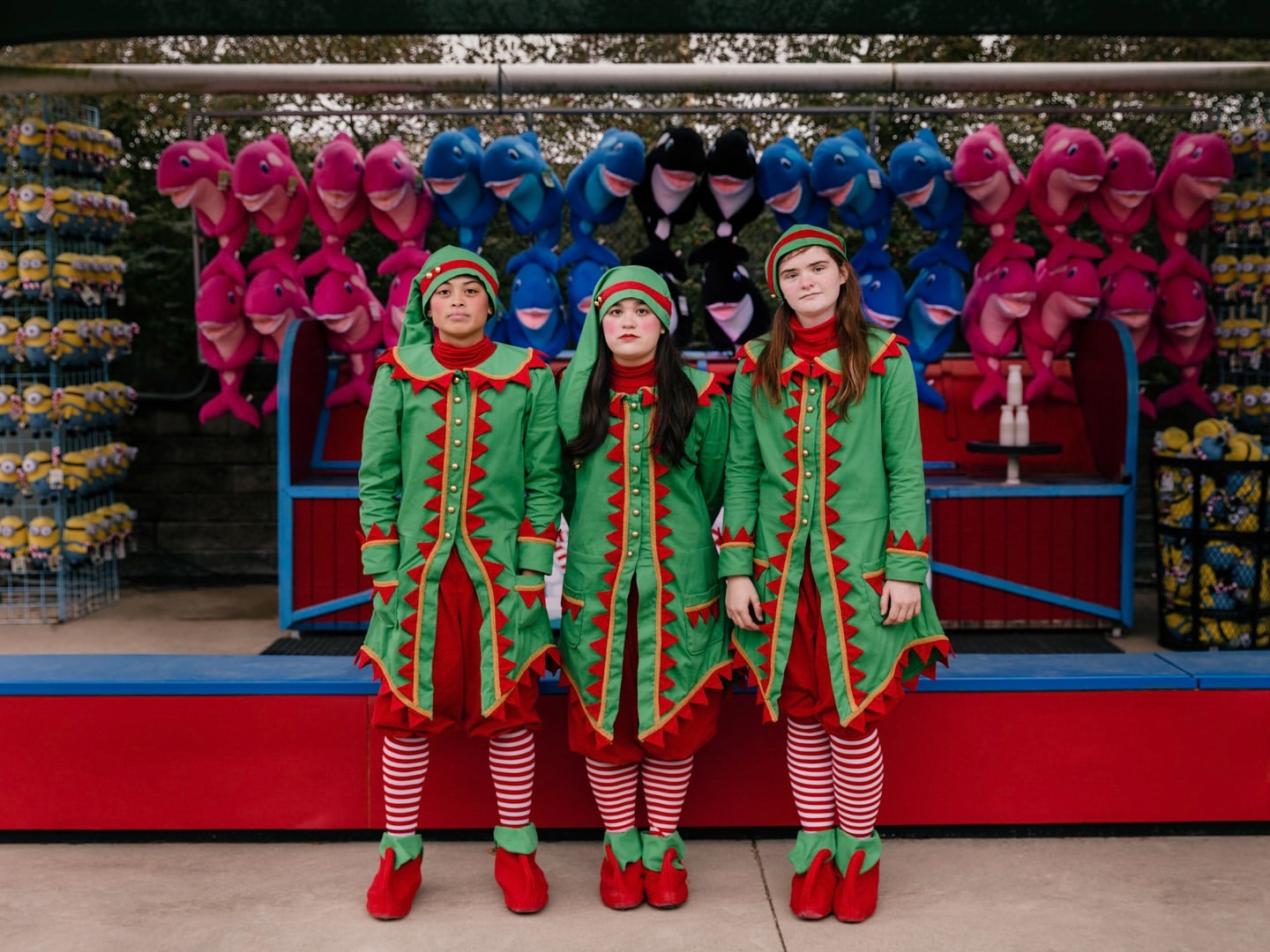 Three people in elf costumes standing in front of a fair booth display with stuffed animal prizes in pink, blue, and yellow.
