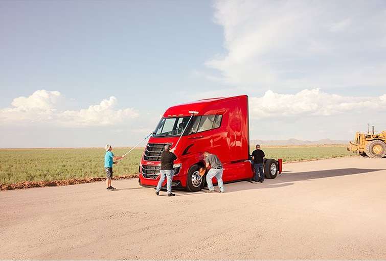 A bright red semi-truck is being washed by a group of people in an open field under a blue sky. Construction equipment is visible.