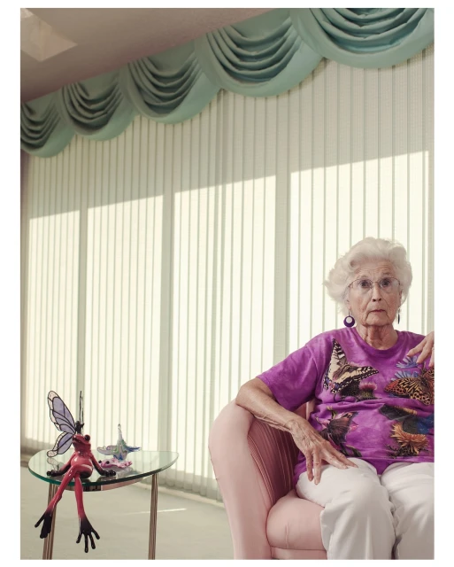 Elderly woman with white hair and glasses, wearing a printed t-shirt, seated in a pink chair next to a table with a frog figurine.