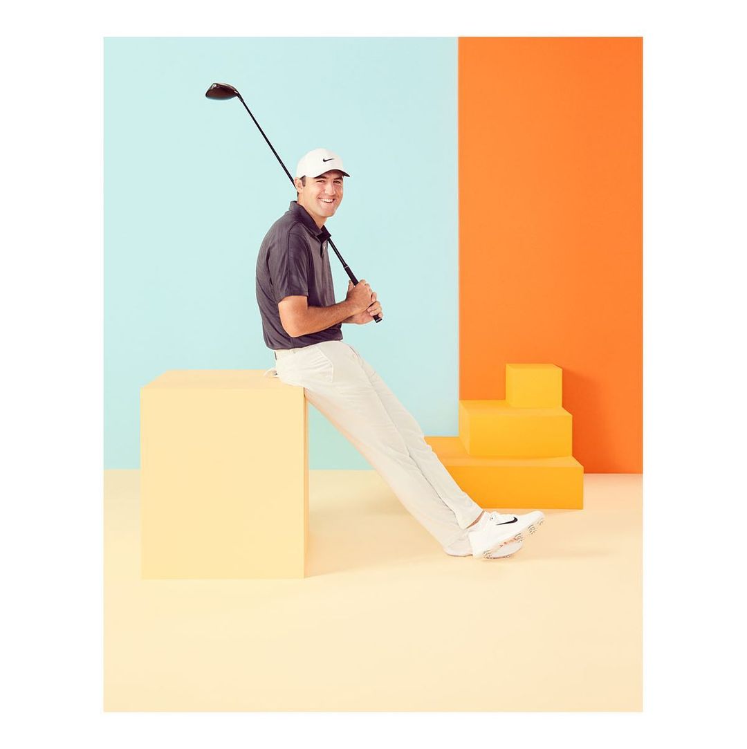 Man in sportswear sits on a yellow cube, holding a golf club against a colorful backdrop of orange and light blue in a studio setting.