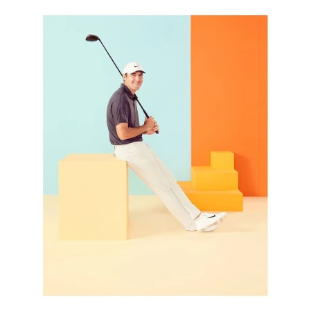 Man in sportswear sits on a yellow cube, holding a golf club against a colorful backdrop of orange and light blue in a studio setting.