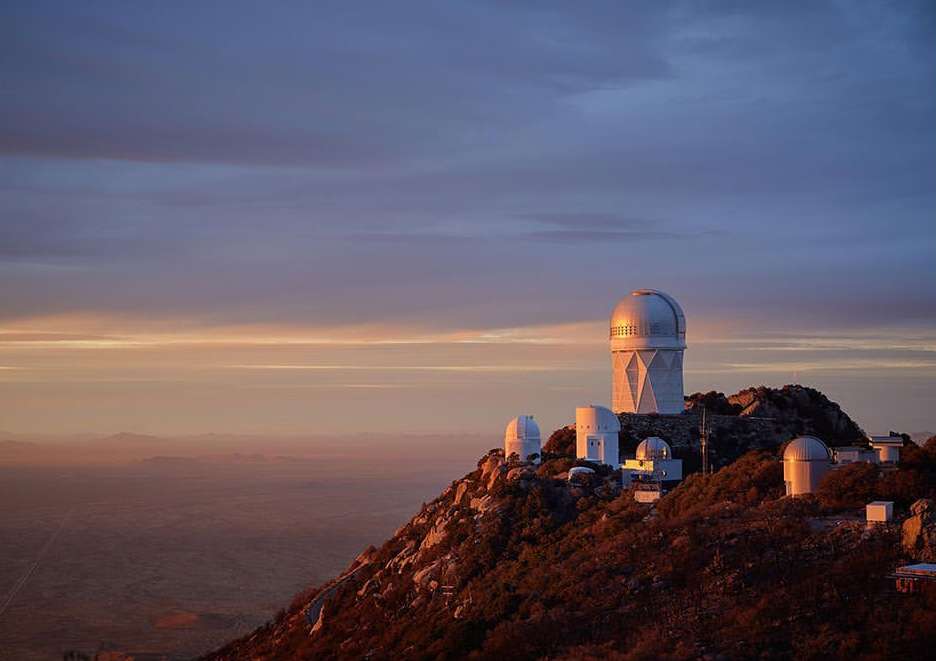 Scenic view of the Kitt Peak Observatory complex on a mountaintop against a sky with soft pastel colors at sunset or sunrise.