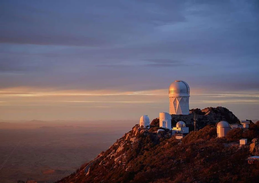 Scenic view of the Kitt Peak Observatory complex on a mountaintop against a sky with soft pastel colors at sunset or sunrise.