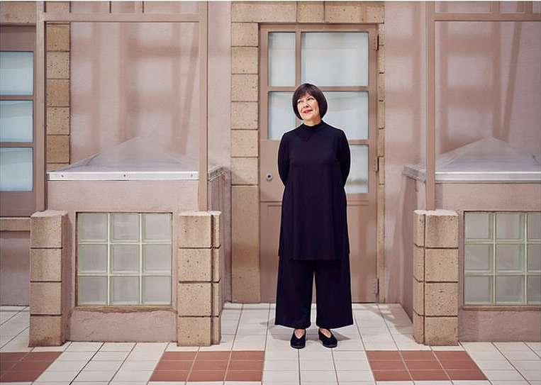 Adult woman in a black outfit stands in front of a pale pink building exterior with tile flooring in a medium, eye-level shot.