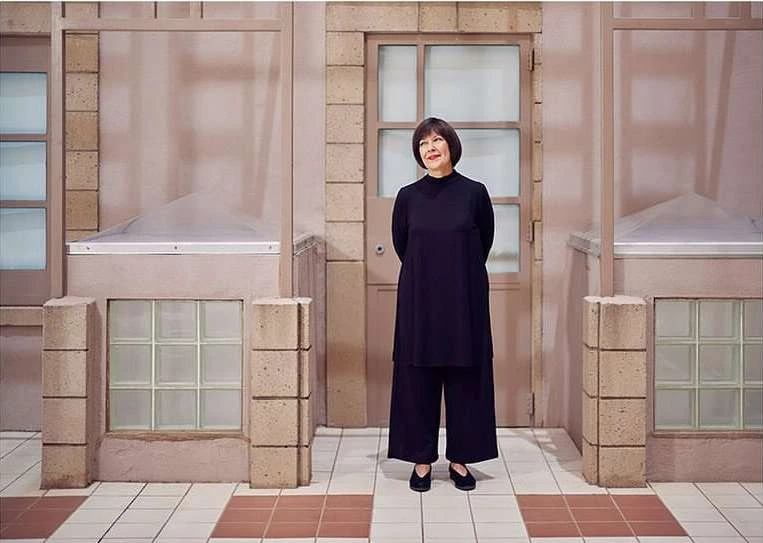 Adult woman in a black outfit stands in front of a pale pink building exterior with tile flooring in a medium, eye-level shot.
