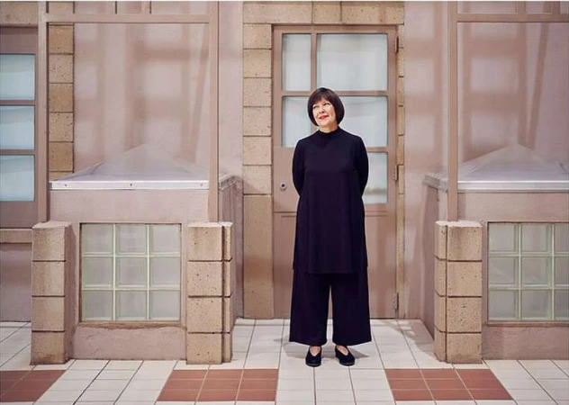Adult woman in a black outfit stands in front of a pale pink building exterior with tile flooring in a medium, eye-level shot.