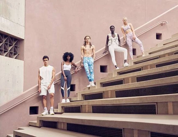 Five diverse models in athletic wear pose on tiered stairs against a light pink wall, creating a fashionable group shot.