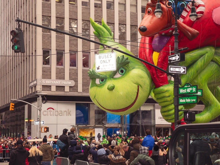 Crowd gathers to watch the Grinch and Max balloon floats at the Macy's Thanksgiving Day Parade in an urban setting with tall buildings.