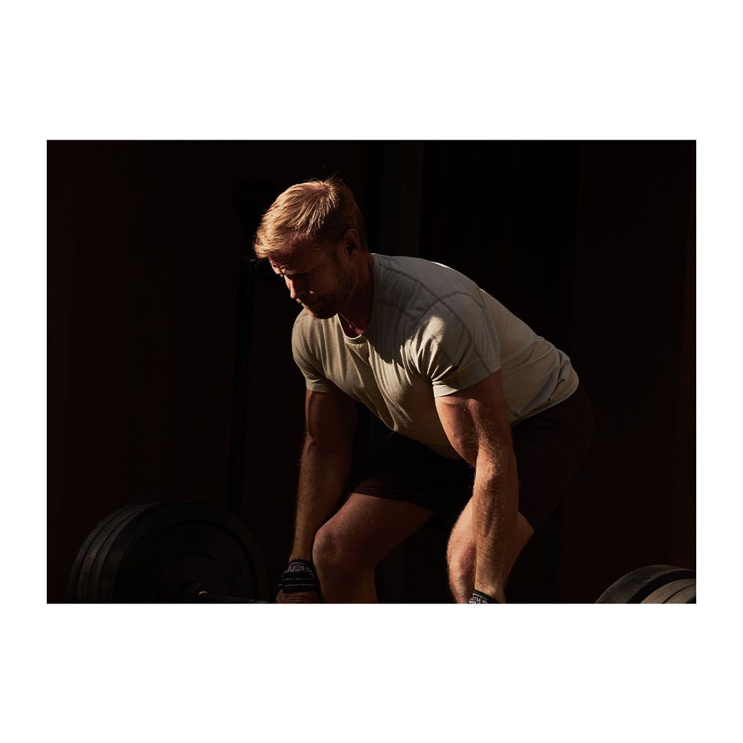 A muscular man prepares for a deadlift in a dimly lit gym, focusing on the weight with a determined expression and strong posture.