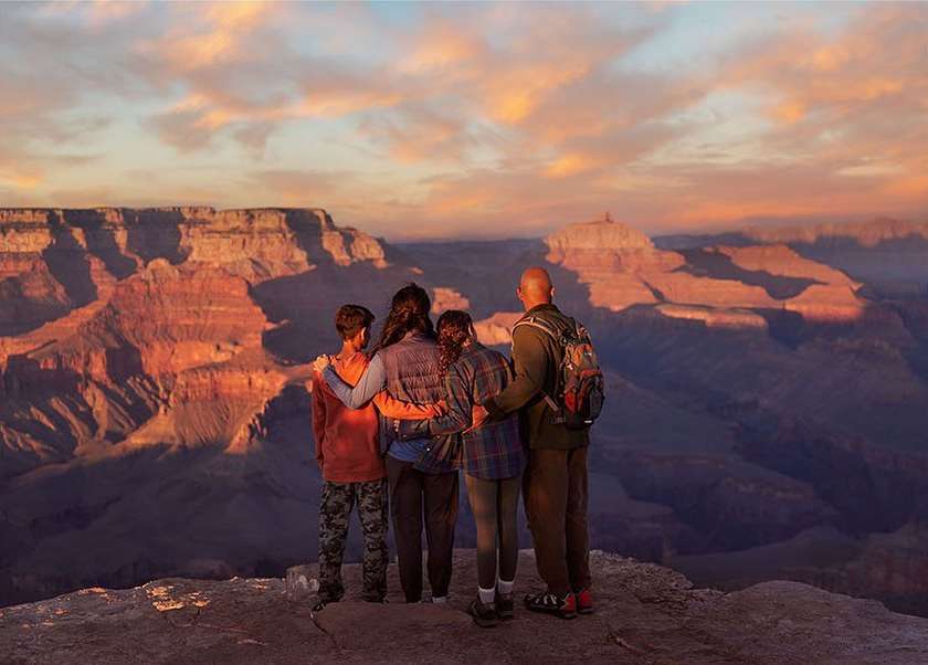 Family of four overlooking the Grand Canyon at sunset. The group is embracing while viewing the vast landscape and colorful sky.