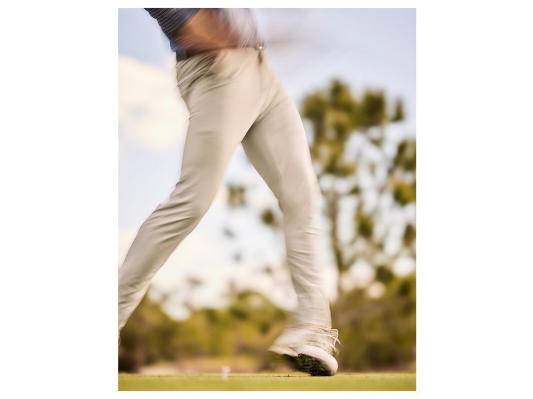 Motion blur shot of an adult male golfer in khaki pants and white golf shoes swinging a club on a sunny day on a golf course.