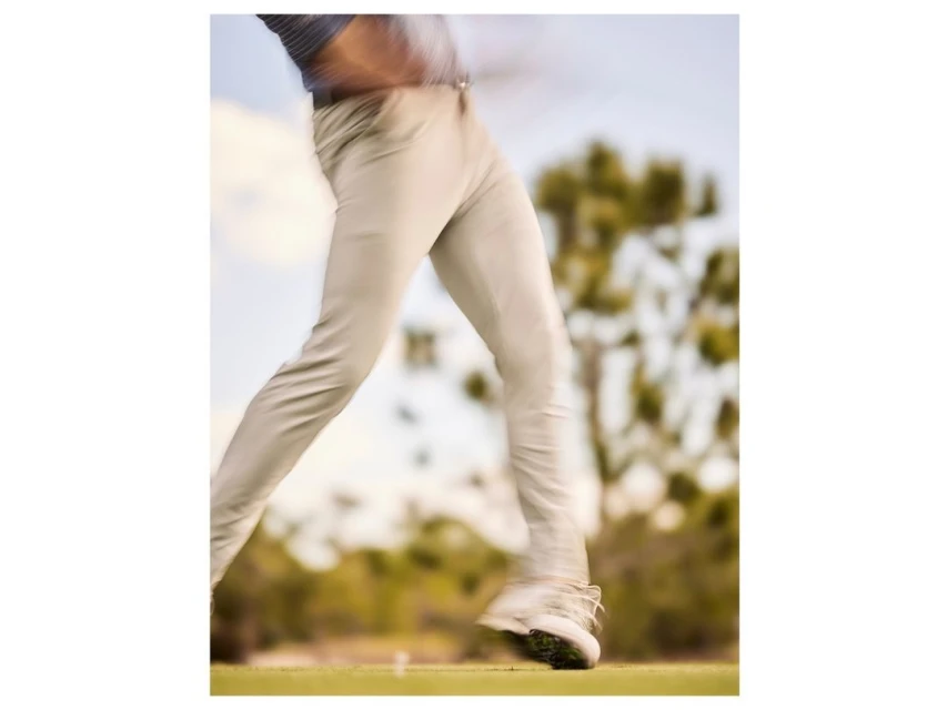 Motion blur shot of an adult male golfer in khaki pants and white golf shoes swinging a club on a sunny day on a golf course.