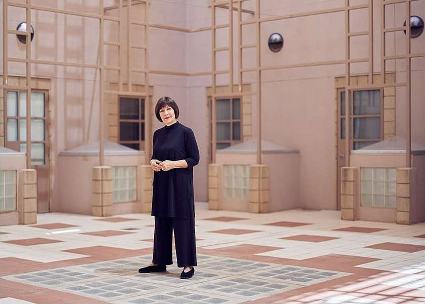 Adult woman in a black outfit stands in an open-air courtyard with a geometric tiled floor, surrounded by pale pink walls with windows.