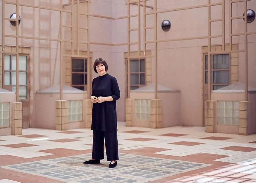Adult woman in a black outfit stands in an open-air courtyard with a geometric tiled floor, surrounded by pale pink walls with windows.