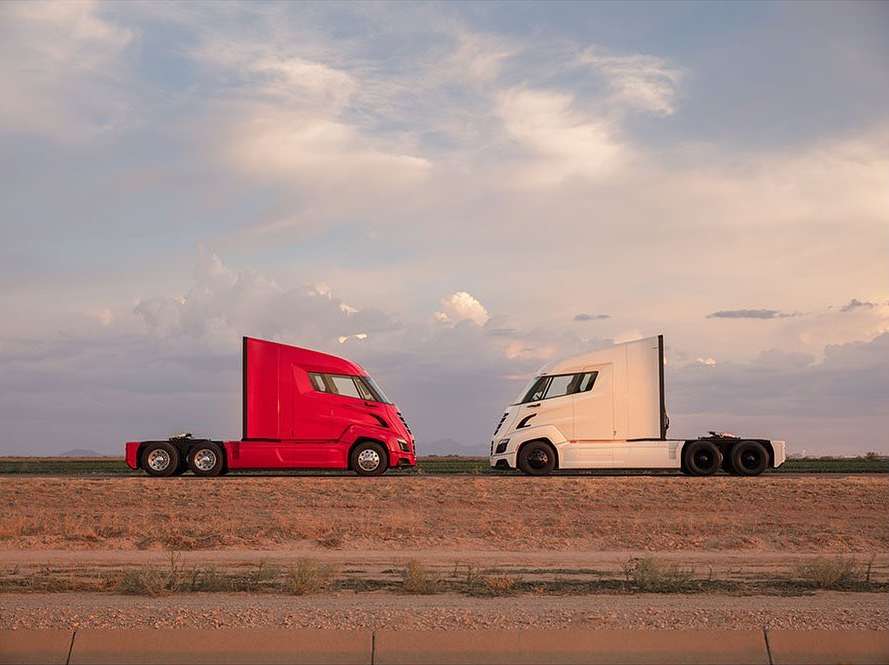 Two semi-trucks, one red and one white, face each other on a rural road under a pastel sunset sky. Automotive industry product shot.
