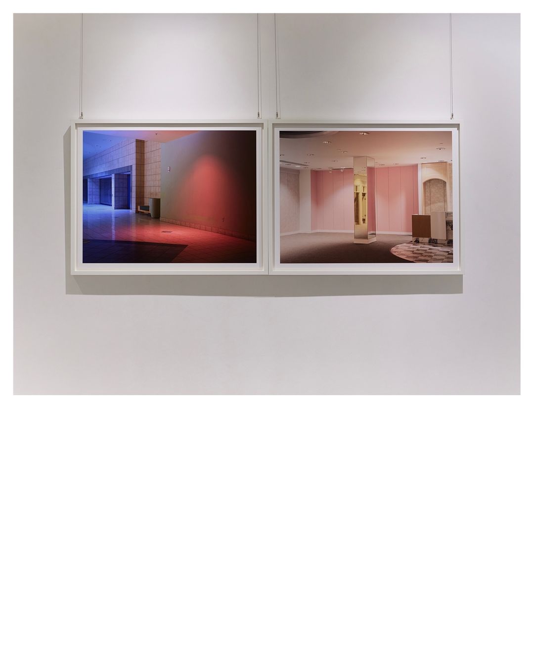 Two framed photographs hang on a white wall, depicting interior spaces illuminated with cool blue and warm pink lighting.
