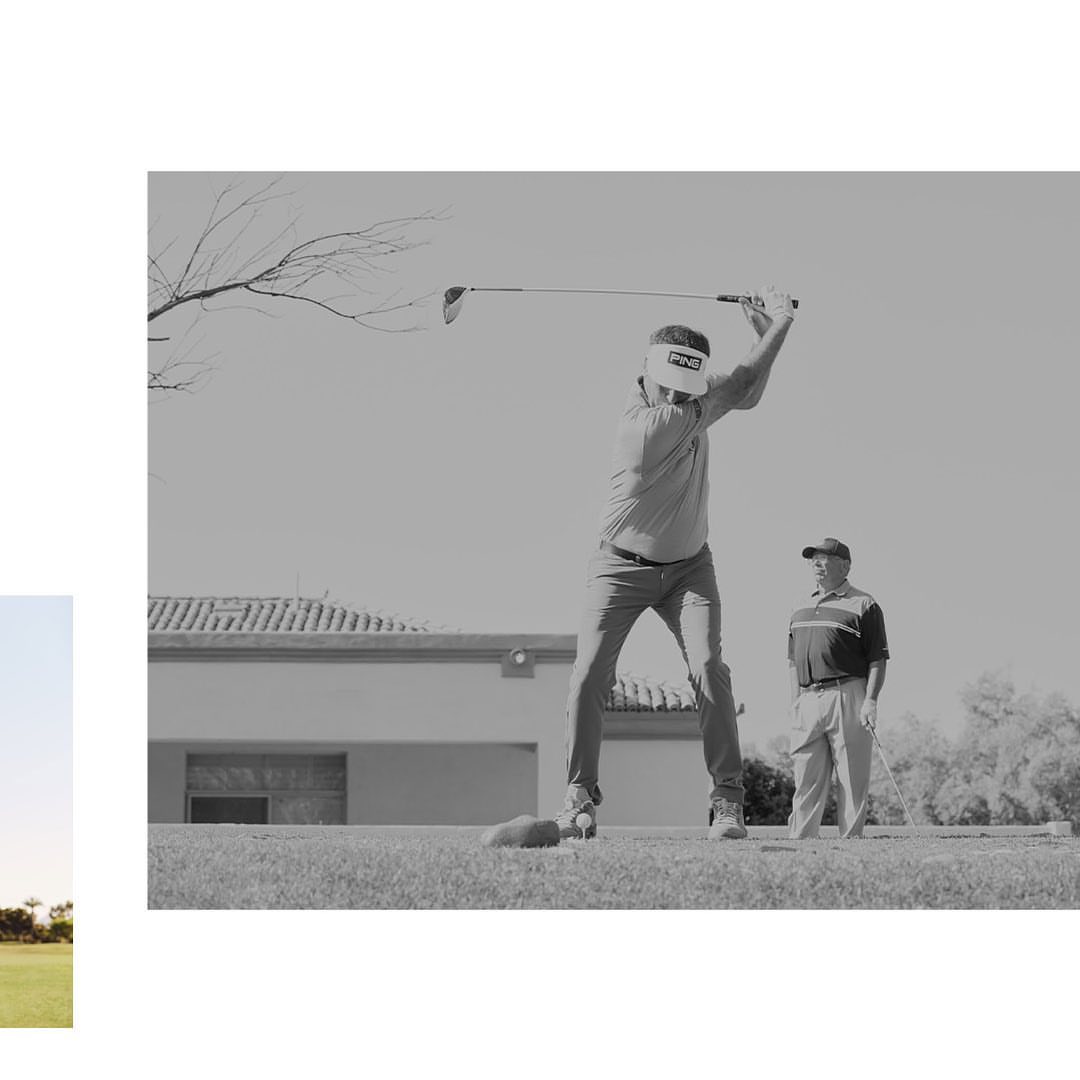 Two adult male golfers on a grassy course, one swinging club, the other watching with golf club in hand, near a building.
