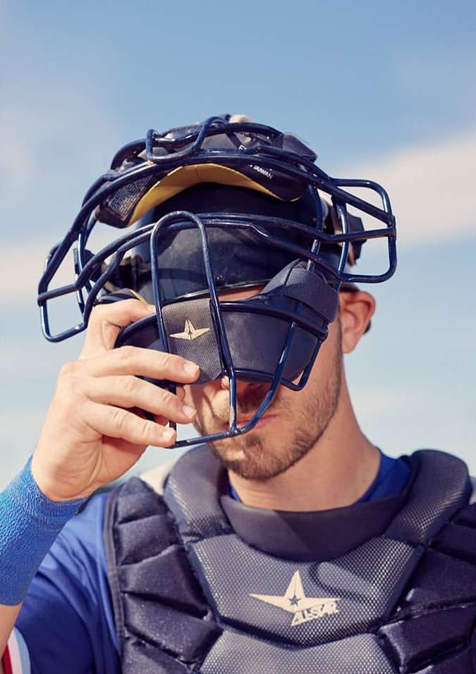 A baseball catcher in full gear adjusts his mask against a light blue sky, creating a portrait focused on the sport and preparation.