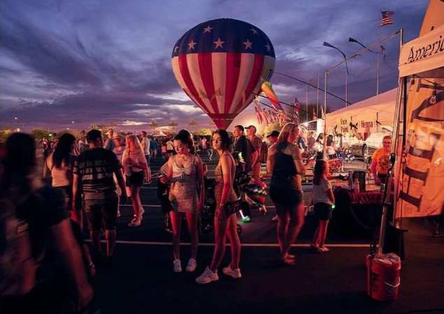 Crowd gathers at an outdoor event with a red, white, and blue hot air balloon in the background under a dusk sky filled with clouds.