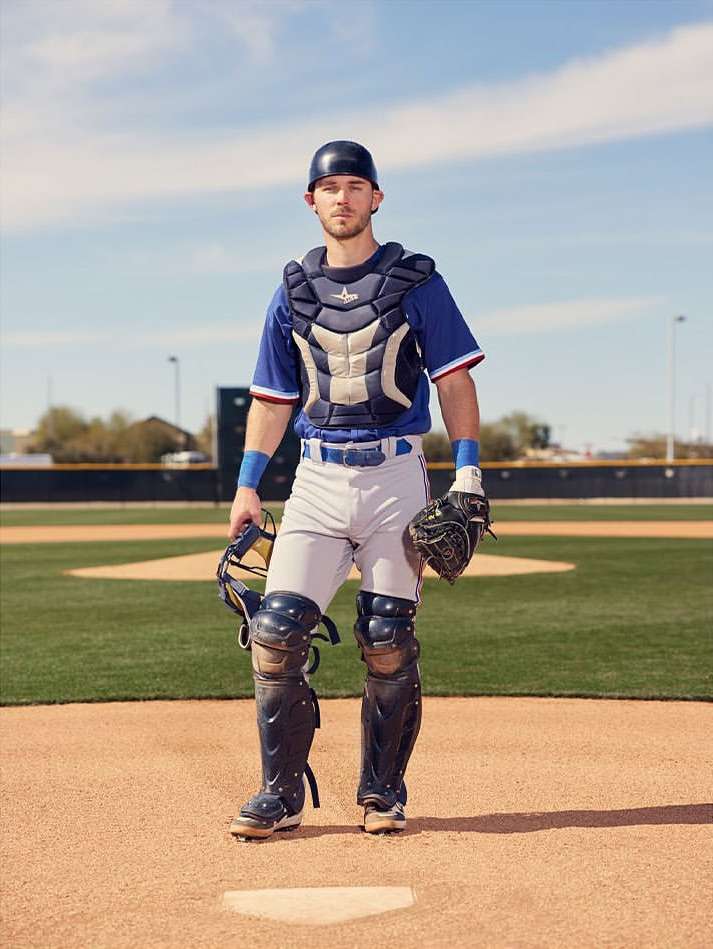 Full-length shot of a male baseball catcher in full gear standing on the baseball field, wearing a blue and white uniform on a sunny day.