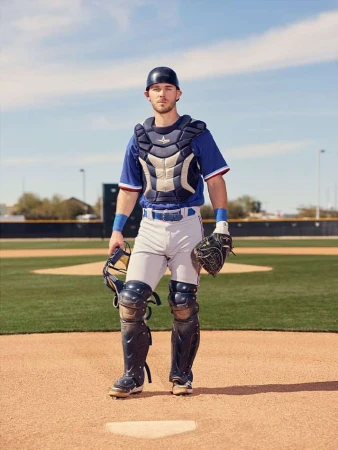 Full-length shot of a male baseball catcher in full gear standing on the baseball field, wearing a blue and white uniform on a sunny day.